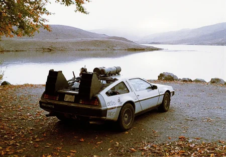 8n8log, retro-styled, heavily modified car parked beside a serene lake surrounded by mountains and grassy hills. The vehicle is a futuristic, sporty design, reminiscent of a DeLorean, with various modifications, including a large rear spoiler, roof-mounted equipment, and visible decals and markings on the body. The car’s exterior shows wear, with scratches and grime, giving it a rugged, utilitarian look, as if it's been used for adventurous journeys. The scene is set in a calm, overcast environment with soft, natural light, enhancing the muted tones of the landscape. Fallen leaves and rocks cover the ground, adding a touch of autumnal decay. The reflective surface of the lake stretches toward distant hills, where the fading horizon blends softly with the pale sky. The car is positioned at an angle, slightly off-center, with its rear facing the viewer, capturing the peaceful yet solitary atmosphere of the environment. film photography aesthetic, film grain effect prominent throughout image, high contrast lighting creating dramatic shadows, grainy film-like texture