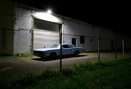 Dilapidated warehouse, nighttime setting, chain-link fence in foreground, overgrown grass. Classic car, metallic blue, parked near fence, low-angle view, dim streetlight overhead casting shadows. Building facade, white, weathered, large garage door, small side door to the right. Isolated urban area, stark lighting, amateurish quality, slightly blurred details, moody atmosphere