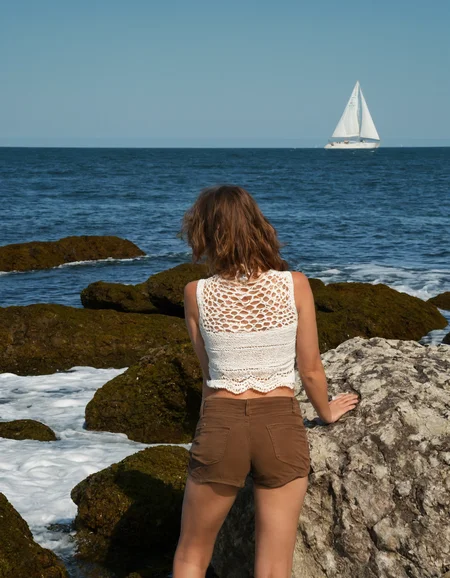 woman, ocean view, standing on rocks, back to camera, white crochet top, brown shorts, waves, algae-covered rocks, horizon, sailboat in distance, blue sky, tranquil scene, outdoor, sunlight, serene, day time, leisure, nature, landscape, wide shot, Fujifilm XT3, Canon R5, Fujicolor Fujichrome Velvia 100