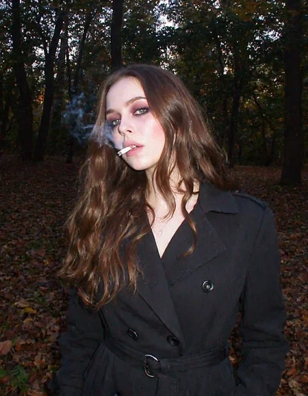 m8vic2, Young woman, early 20s, casual style, wearing dark trench coat, long wavy brown hair cascading over her shoulders, looking direct at the viewer, gothic heavy makeup, outdoors in the dark forest, autumn, lit cigarette between her lips, smoke comes from cigarette, sunlight filtering through the trees