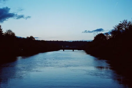 8n8log, Serene river landscape, calm blue water reflecting light, distant bridge crossing waterway, dense tree lines on both riverbanks, silhouetted dark trees creating contrast, clear light blue sky with few scattered clouds, dusk or early evening atmosphere, forested horizon line, distant mountains or hills barely visible, peaceful rural or suburban setting, faint lights visible in distance, no people present, natural outdoor environment, cool color palette dominated by blues and dark silhouettes, slight film grain texture, slightly underexposed image quality, tranquil water surface with subtle ripples, film photography aesthetic