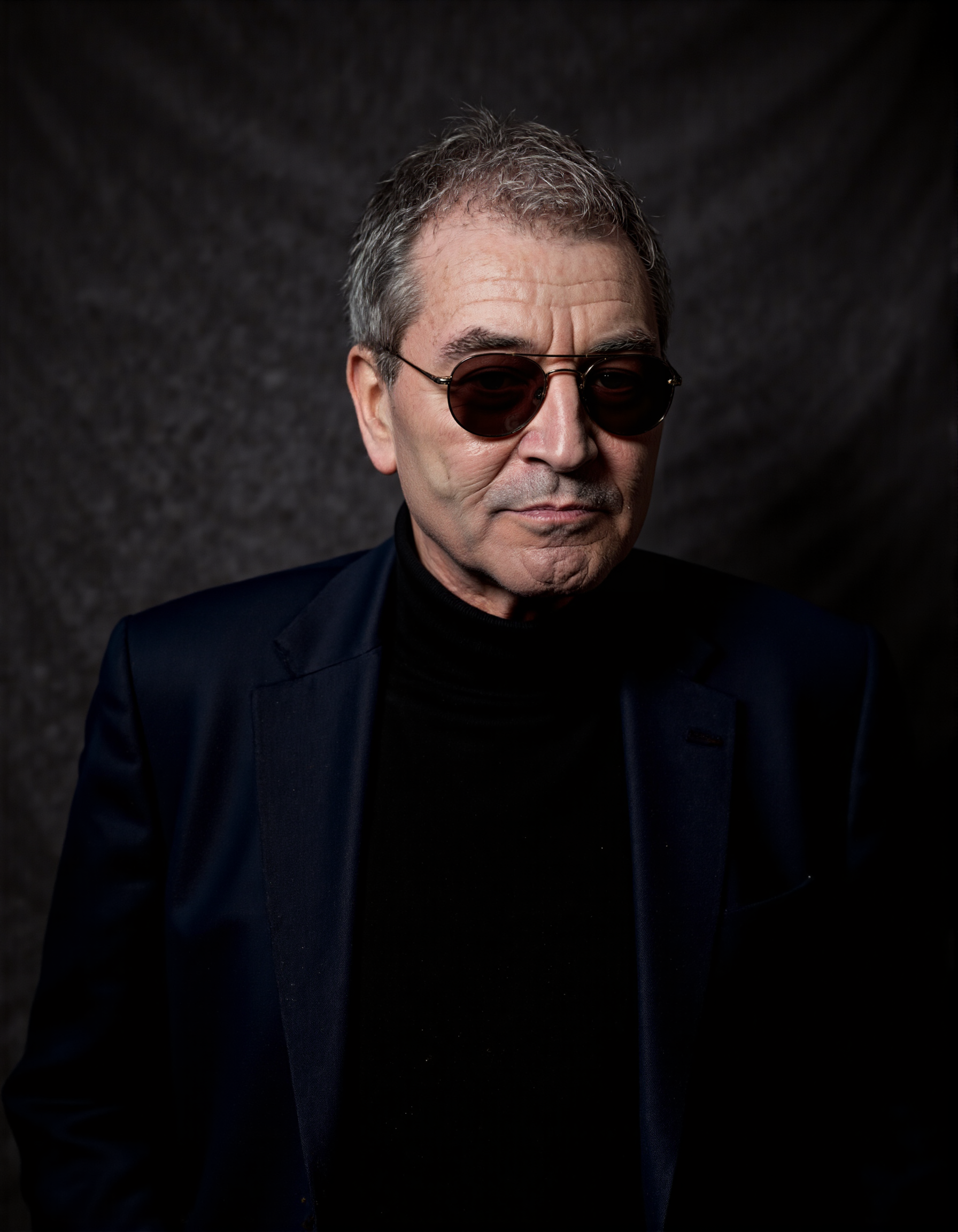 <lora:IAN_GILLAN:1>, The image is a high-resolution photograph featuring an elderly man with a distinguished appearance. He has short, graying hair and wears round, dark-tinted sunglasses that obscure his eyes. His skin is fair with visible wrinkles, indicating his age. He is dressed in a dark navy blue blazer over a black turtleneck sweater, which contrasts with his light skin tone. The background is a plain, dark gray, almost black, textured fabric that provides a minimalist backdrop, focusing attention solely on the subject. The lighting is soft and even, highlighting the textures of his clothes and the subtle shadows on his face. The photograph captures him from the chest up, with his head slightly tilted to the right, giving him a contemplative and introspective look. The overall mood of the photograph is serious and thoughtful, enhanced by the man's expression and the simplicity of the background.
