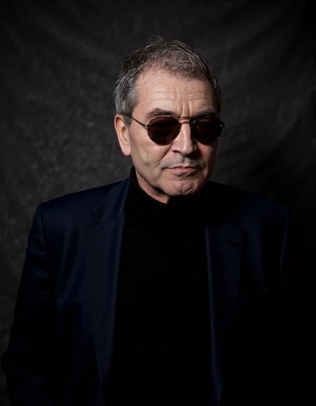 <lora:IAN_GILLAN:1>, The image is a high-resolution photograph featuring an elderly man with a distinguished appearance. He has short, graying hair and wears round, dark-tinted sunglasses that obscure his eyes. His skin is fair with visible wrinkles, indicating his age. He is dressed in a dark navy blue blazer over a black turtleneck sweater, which contrasts with his light skin tone. The background is a plain, dark gray, almost black, textured fabric that provides a minimalist backdrop, focusing attention solely on the subject. The lighting is soft and even, highlighting the textures of his clothes and the subtle shadows on his face. The photograph captures him from the chest up, with his head slightly tilted to the right, giving him a contemplative and introspective look. The overall mood of the photograph is serious and thoughtful, enhanced by the man's expression and the simplicity of the background.