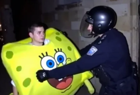 Police officer in riot gear, mid-30s, wearing helmet with visor, dark uniform with visible badge number, padded gloves, in the act of detaining a young man dressed as a sponge bob character. The young man, early 20s, Caucasian, short dark hair, wearing a bright, oversized yellow costume resembling a sea sponge, with a wide-eyed, cartoonish expression printed on it, standing pose, looking forward. Outdoor setting, dimly lit, stone wall in background, image slightly blurred, amateurish quality.,