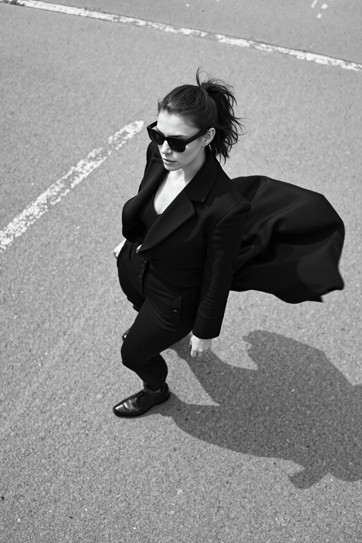 sony_a7III professional style photo, Black and white photo of a stylish woman, early 20s, medium build, ponytail haircut, wearing angular black sunglasses and a flowing dark coat, mid-twirl creating dynamic motion in the fabric, standing on a textured concrete surface with faint painted lines, strong shadows and light interplay casting dramatic patterns, high contrast lighting accentuating the minimalist and cinematic mood, overhead perspective enhancing depth and movement in the composition