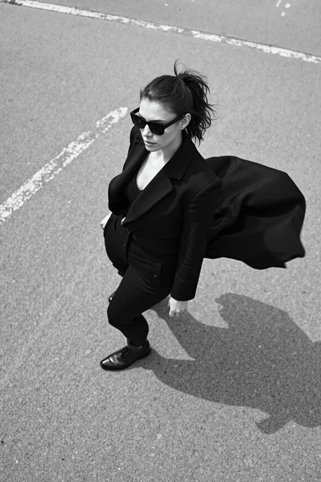 sony_a7III professional style photo, Black and white photo of a stylish woman, early 20s, medium build, ponytail haircut, wearing angular black sunglasses and a flowing dark coat, mid-twirl creating dynamic motion in the fabric, standing on a textured concrete surface with faint painted lines, strong shadows and light interplay casting dramatic patterns, high contrast lighting accentuating the minimalist and cinematic mood, overhead perspective enhancing depth and movement in the composition