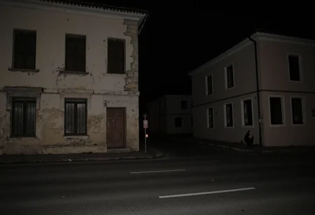 Dark, nighttime street scene, two shadowed buildings, left building has aged plaster, closed wooden shutters, bars on windows, right building with a monochrome facade, windows with no visible details, silhouette of a rooster painted on right building wall, asphalt road in foreground, minimal street lighting, no people or vehicles, quiet, eerie atmosphere, slightly underexposed, amateurish quality.,