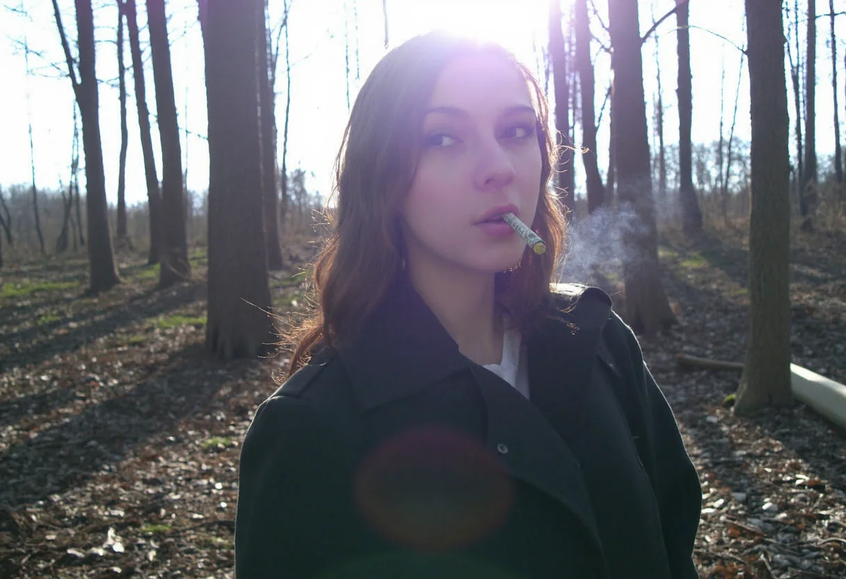 low-resolution photo, shot on a mobile phone, noticeable graininess, no visible JPEG artifacts, some lens flare from the sunlight in the background. Young woman, early 20s, casual style, wearing dark trench coat, long wavy brown hair cascading over her shoulders, looking direct at the viewer, makeup, outdoors in the dark forest, autumn, holding a cigarette to her lips, smoking cigarette, sunlight filtering through the trees