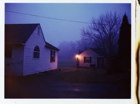 p0l2rd, Residential neighborhood at dusk, foggy evening setting, two houses visible, left house larger, white exterior, arched window, dark roof, right house smaller, grey exterior, black shutters, streetlamp glowing orange on the left side, bare trees silhouetted in the background, dark mailbox, empty driveway, dim lighting conditions, purple-blue haze dominates, outdoor urban environment, low visibility, street and grass barely discernible, overcast sky.  prominent film grain, high contrast lighting casting dramatic shadows, grainy film-like texture,  polaroid-style format with white border, distinctive burnt edge, photograph appears aged or partially developed, with the right side fading into white