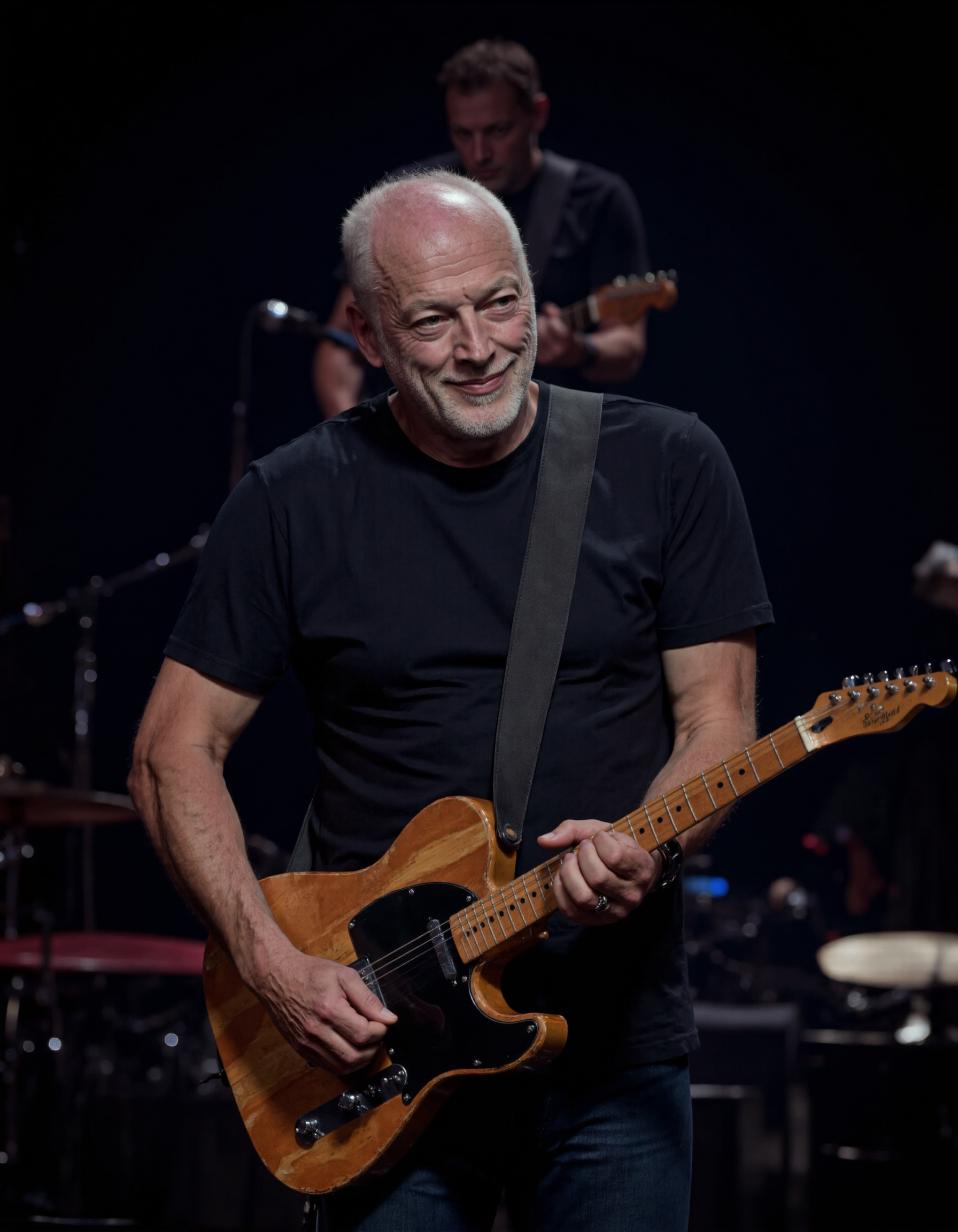 <lora:DAVID_GILMOUR:1>, The image is a photograph featuring a middle-aged man with a bald head and a short white beard. He is captured mid-song, with a slight smile and a relaxed expression. The man is wearing a plain, dark-colored t-shirt and has a strap of a guitar slung over his shoulder. The guitar, which he is holding, is a wooden electric guitar with a sleek finish and visible frets. The background is dark and out of focus, likely indicating that the photograph was taken at a live concert or performance setting. The lighting is focused on the subject, highlighting his face and the guitar, while the rest of the scene remains dim. The man's eyes are looking slightly to his left, adding a sense of depth and engagement to the image. The photograph captures the moment of performance, emphasizing the musician's connection with his instrument and the audience. The overall mood is one of relaxed, joyful engagement with the music.