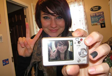 aesthetic2000s, amateur photo, Canon PowerShot A620, compact camera, retro camera, digital screen, female hands, nail polish, Hello Kitty stickers, selfie, young goth woman, mirror reflection, V pose, smile, bedroom, curtain, wall fan, nameplate, warm lighting, close-up, handheld, indoors, photograph, nostalgia, casual setting