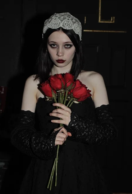 Young woman, late teens to early 20s, pale complexion, dark straight shoulder-length hair, wearing a vintage-inspired black lace dress, dramatic dark eyeliner, pale foundation, deep red lipstick, white lace headpiece, standing in a mysterious dark setting, holding a bouquet of wilted red roses close to her chest, ethereal goth style, eerie atmosphere, image appears slightly overexposed, hinting at an amateurish quality, low lighting