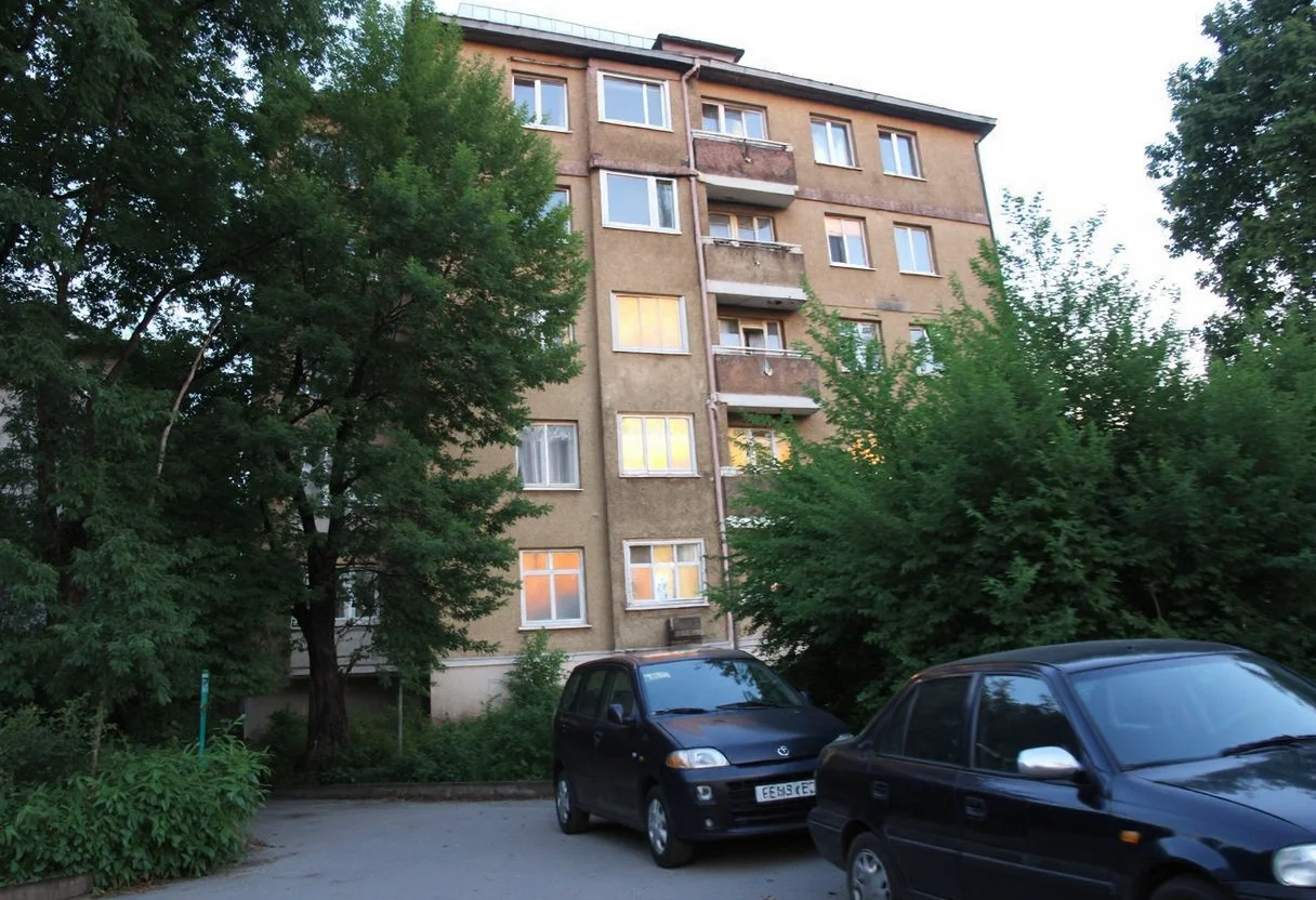 A slightly faded, multi-story Soviet-era apartment building stands with a worn, light brown fa ade, multiple windows with glowing warm lights indicating varying activities indoors. The structure is partly obscured by lush, green trees on the left and right, blending with the urban environment. Two parked cars, a dark sedan and a compact black vehicle, can be seen in the foreground in a narrow alleyway, bordered by untamed shrubbery. The image lacks sharpness, with an overall amateurish quality, and depicts a serene, early evening atmosphere as twilight descends, with subtle blurred details of the foliage