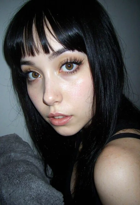 d1g1cam, amateur photo, low-lit, overexposure, Low-resolution photo, shot on a mobile phone, noticeable noise in dark areas, visible JPEG artifacts, dynamic. Young woman, early 20s, mixed Asian-European descent, intense gaze directly at camera, striking hazel eyes, dramatic false eyelashes, defined thick dark eyebrows, short straight bangs, long black hair, natural freckles across nose, matte nude lipstick, sharp facial features, black top visible at neckline, gray plush fabric visible in corner, close-up selfie angle, indoor setting, soft natural lighting from side, smooth skin texture, neutral background, portrait orientation photo