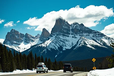 sony_a7III professional style photo, Snow-capped mountain with rugged peaks, large mass of rock and ice, forested slopes with dense evergreen trees, two vehicles on winding road, white SUV with roof cargo, black SUV trailing, clear blue sky with scattered white clouds, deer crossing sign on the right side, natural outdoor setting, bright sunlight, daytime, expansive view from low angle, crisp and clear visibility, vibrant colors, deep green forest contrasting with white snow, background type mountain landscape, lighting natural