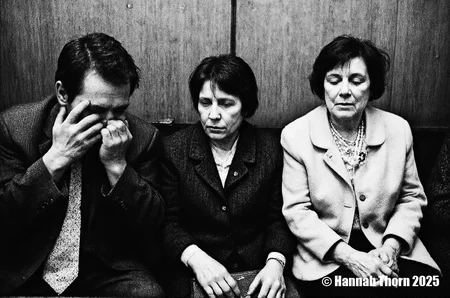A black and white photograph from the front about a man in a suit sitting on the left side of the image, covering his face with his hands, with a harmonica in his mouth, in an elevator with a wooden wall in the background. the image also shows two women, one older and one younger, sitting next to each other, both with expressions of distress on their faces. the older woman, on the right side, appears to be a woman with short hair, wearing a beige jacket and a pearl necklace, and the younger woman, also with short black hair, is also wearing a black jacket. the elderly woman, who is looking down at the older one, has a serious expression on her face, while the younger one is looking up at her with a concerned expression.<lora:_Flux\Anders_Petersen.safetensors:0.9:0.9>