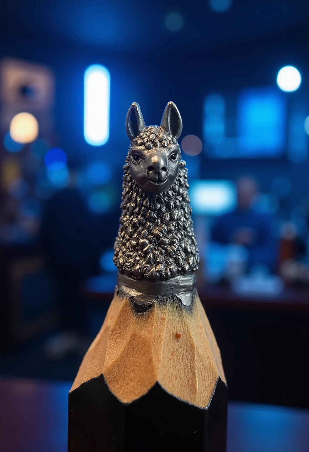 Mini sculpture on the tip of the pencil, showing a llama head potrait. The background is a night bar with blue lights.