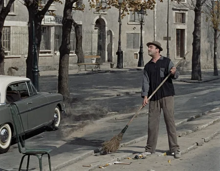 A movie still of a man holding a broom and standing on a street. A car is parked on the street, and a chair can be seen close to the bench., ground vehicle, outdoors, car, motor vehicle, tree, hat, building, broom, pants, black hair, holding, scenery, black shirt, long sleeves, road, lamppost, day, bench, standing