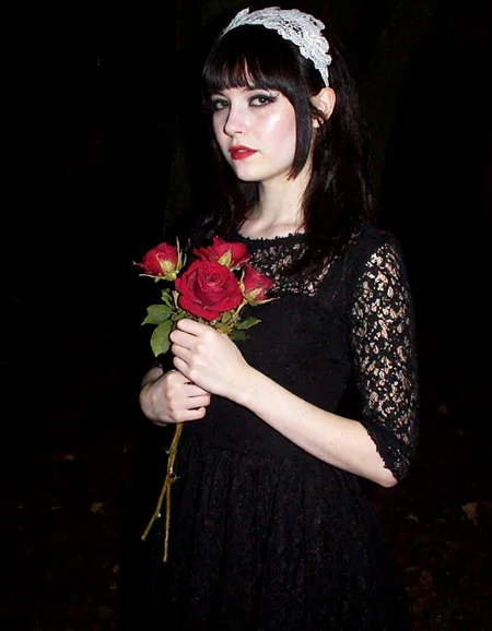 m8vic2, Young woman, late teens to early 20s, pale complexion, dark straight shoulder-length hair, wearing a vintage-inspired black lace dress, dramatic dark eyeliner, pale foundation, deep red lipstick, white lace headpiece, standing in a mysterious dark setting, holding a bouquet of wilted red roses close to her chest, ethereal goth style, eerie atmosphere,