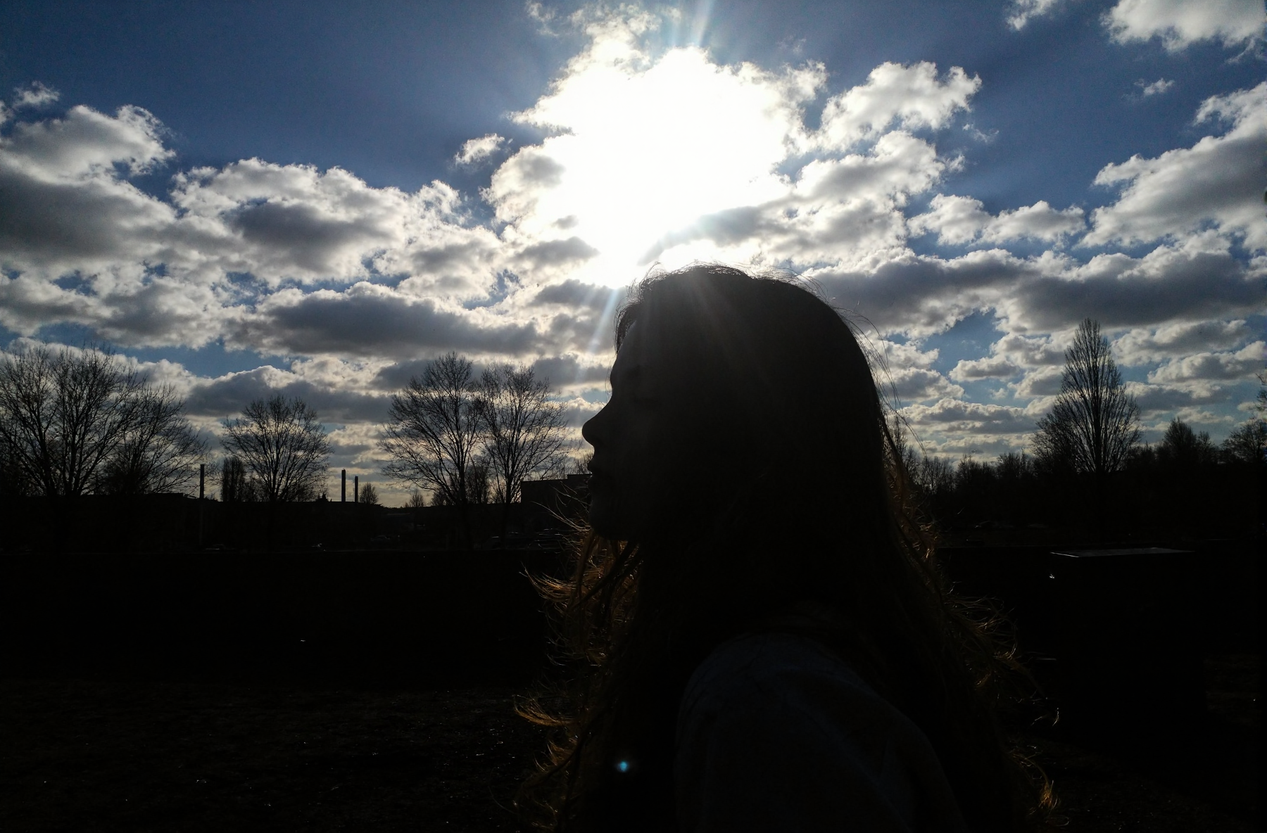 l3n0v0. portrait of teenager girl, flowing tousled hair, from side view angle, looking at the viewer, dreamy expression, closed eyes, bare winter trees, dramatic cloudy sky, bright sunburst through clouds, deep blue sky patches, industrial urban setting, dark shadowed foreground, late afternoon lighting, silhouetted landscape, underexposed ground elements, natural dramatic lighting, high contrast image. amateur quality, candid style
