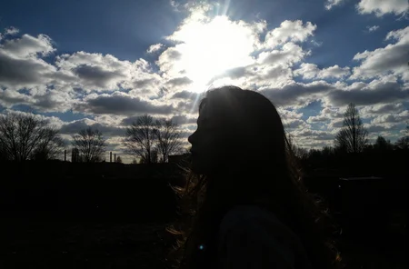 l3n0v0. portrait of teenager girl, flowing tousled hair, from side view angle, looking at the viewer, dreamy expression, closed eyes, bare winter trees, dramatic cloudy sky, bright sunburst through clouds, deep blue sky patches, industrial urban setting, dark shadowed foreground, late afternoon lighting, silhouetted landscape, underexposed ground elements, natural dramatic lighting, high contrast image. amateur quality, candid style
