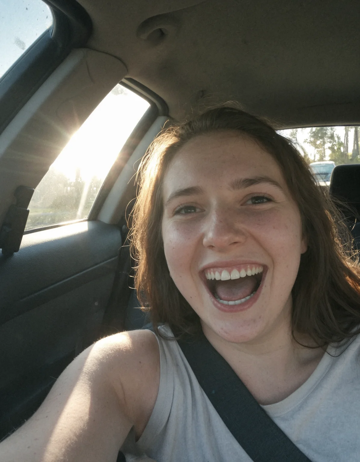Authentic share, Spontaneous, authentic, natural light: Car selfie. Person in driver's seat, light coming through windshield. Seatbelt visible, dashboard and steering wheel in frame. Subject possibly caught mid-laugh or making a face.