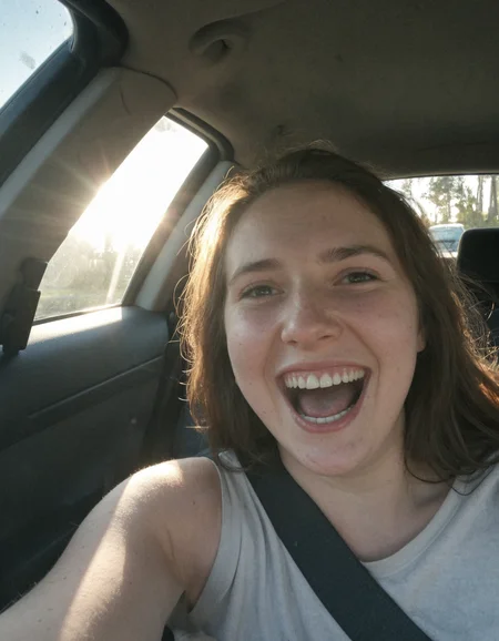 Authentic share, Spontaneous, authentic, natural light: Car selfie. Person in driver's seat, light coming through windshield. Seatbelt visible, dashboard and steering wheel in frame. Subject possibly caught mid-laugh or making a face.