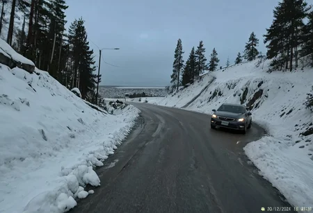 shit_cm_shot, photograph, winter road, snow-covered, icy cliff, pine trees, overcast sky, car driving away, left side, right side rocky cliff, white snow, gray sky, blue hour lighting, leading lines, symmetry, cold, serene, peaceful, nature, wilderness, remote location, high angle shot, slight fisheye effect, timestamp in bottom right corner