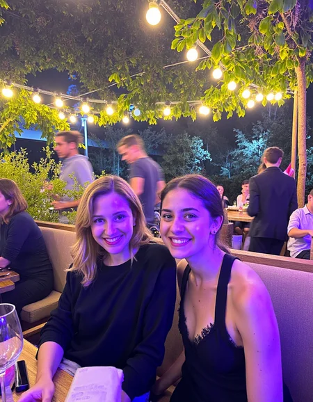 In a candid medium shot taken at an outdoor café during the evening, two young women are seated close together, smiling at the camera amidst a lively social setting. The color palette is composed of warm yellow lights and dark shadows, creating a cozy and intimate atmosphere. The woman on the left has long blonde hair, loosely styled, and wears a black top, while the woman on the right has her dark hair pulled back and wears a black sleeveless top. The lighting is ambient, coming from strings of lights hanging overhead and casting a warm glow over the subjects. There is noticeable grain due to the low-light condition, and some mild motion blur from the other guests moving around in the background, which slightly detracts from the clarity of the image. The setting features lush green foliage above, strings of glowing bulbs, and blurred figures of other patrons, contributing to the bustling, relaxed evening environment <lora:amateurphoto-v5-14-15-1-1:0.45>
