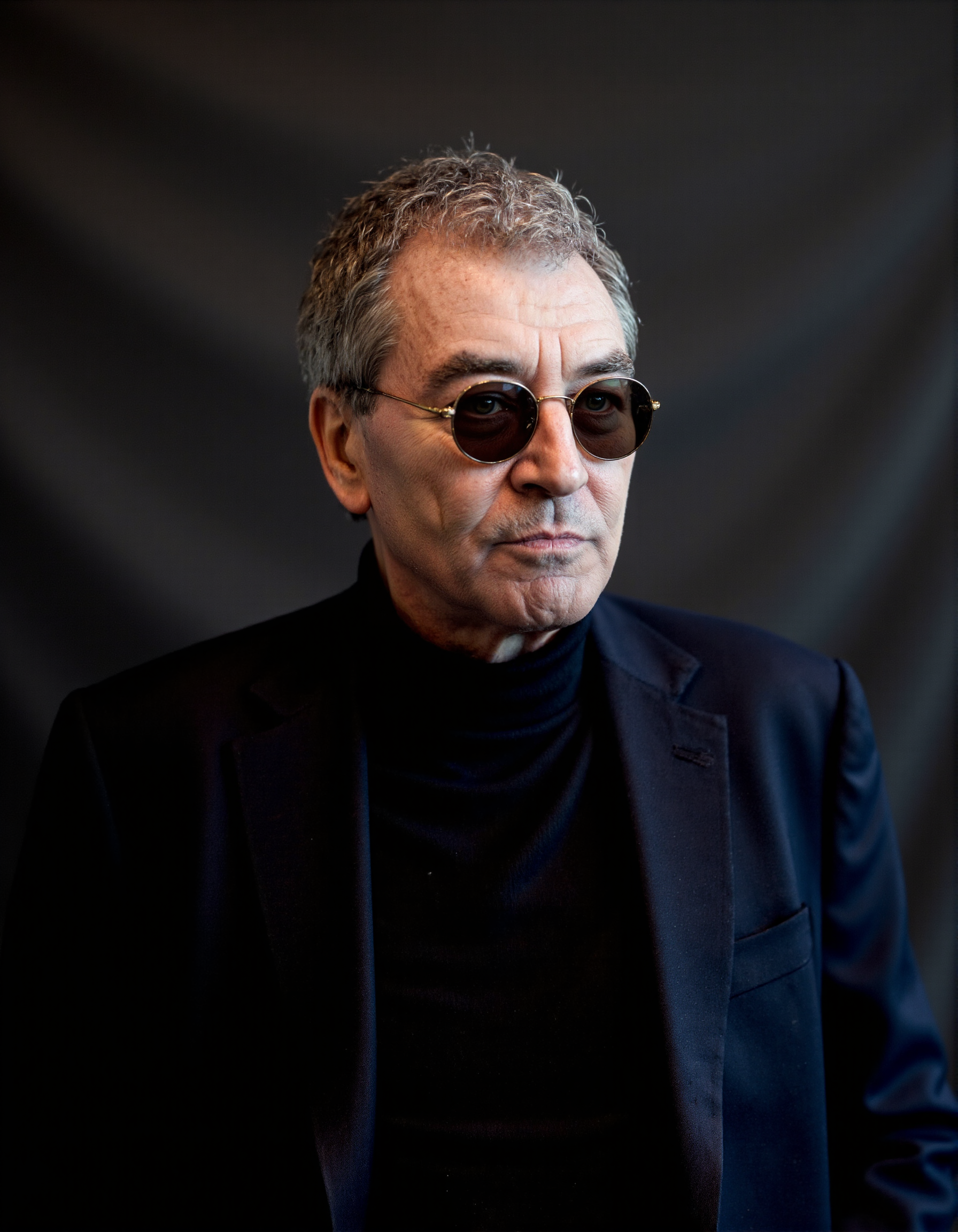 <lora:IAN_GILLAN:1>, The image is a high-resolution photograph featuring an elderly man with a distinguished appearance. He has short, graying hair and wears round, dark-tinted sunglasses that obscure his eyes. His skin is fair with visible wrinkles, indicating his age. He is dressed in a dark navy blue blazer over a black turtleneck sweater, which contrasts with his light skin tone. The background is a plain, dark gray, almost black, textured fabric that provides a minimalist backdrop, focusing attention solely on the subject. The lighting is soft and even, highlighting the textures of his clothes and the subtle shadows on his face. The photograph captures him from the chest up, with his head slightly tilted to the right, giving him a contemplative and introspective look. The overall mood of the photograph is serious and thoughtful, enhanced by the man's expression and the simplicity of the background.