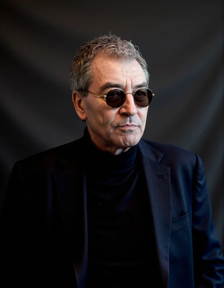 <lora:IAN_GILLAN:1>, The image is a high-resolution photograph featuring an elderly man with a distinguished appearance. He has short, graying hair and wears round, dark-tinted sunglasses that obscure his eyes. His skin is fair with visible wrinkles, indicating his age. He is dressed in a dark navy blue blazer over a black turtleneck sweater, which contrasts with his light skin tone. The background is a plain, dark gray, almost black, textured fabric that provides a minimalist backdrop, focusing attention solely on the subject. The lighting is soft and even, highlighting the textures of his clothes and the subtle shadows on his face. The photograph captures him from the chest up, with his head slightly tilted to the right, giving him a contemplative and introspective look. The overall mood of the photograph is serious and thoughtful, enhanced by the man's expression and the simplicity of the background.