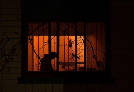 The image shows a dimly lit scene through a window with decorative iron bars. Inside, a silhouette of a person is visible against an orange-hued background, softly illuminated by an overhead light. The person appears to be standing or sitting near a table, with their head slightly bent forward, obscuring detailed features and making it difficult to determine age or race. The building's exterior is clad in a grid-patterned facade, and bare branches partially frame the window, adding an element of starkness. amateur quality, low resolution, low-light conditions and external perspective
