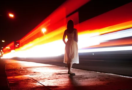 night-time urban scene featuring a lone woman standing still in the center of the frame, with vibrant red, orange, and white light trails swirling around her, captured with long exposure to create a sense of motion and energy. The woman, dressed in a flowing dress that moves slightly with the breeze, is illuminated by the surrounding light, her silhouette contrasting against the dark night. The ground beneath her is wet, reflecting the bright lights and adding depth to the scene. The light trails, possibly from passing cars or other fast-moving vehicles, streak across the image in curved lines, wrapping around the woman and adding a surreal, almost futuristic feel to the composition. The lighting is moody, with a rich contrast between the dark background and the bright, colorful light streaks. The overall atmosphere is one of stillness amidst chaos, blending human presence with the fast-paced movement of the city