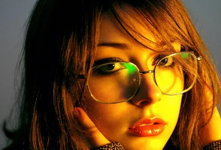 extreme close-up portrait of a teen girl, Eastern European woman, light skin, styled with crimped hair cascading down her shoulders, adding playful texture and volume, subtle center-parted bangs framing her face. She is wearing thin, rectangular metal-framed glasses typical of early 2000s fashion, glossy nude lips, lightly flushed cheeks, and thin, arched eyebrows that emphasize her delicate facial features. Her expression is self-assured and composed, with her gaze slightly off-camera. bright soft lighting highlighting her facial features, glasses, and textured hair, capturing the vibrant and stylish essence of early 2000s trends. strands falling across face, slight off-center position to the left of the frame, body slightly turned toward viewer, golden lighting emphasizing facial features, soft glow around eyes and cheekbones, subtle shadows on parts of face, dark-grey out-of-focus background, soft intimate melancholic atmosphere