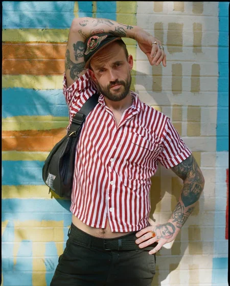 A man with a beard and tattoos standing in front of a colorful striped wall, posing confidently with one hand on his hip and the other resting on his thigh. he is wearing a red and white striped button-up shirt, black pants, and a black bag slung over his shoulder. his body is slim, and he has a beard. he has several tattoos on his arms, including one on his left arm and another on his right arm. he also has a cap on his head and a ring on his finger. the man is looking directly at the camera with a serious expression. the background is a vibrant outdoor setting with a striped wall in shades of blue, green, and yellow.
ðï¸ [Inpainted with Loras]: flux1-depth-dev-lora
ð¯<lora:flux\loratools\flux1-depth-dev-lora.safetensors:0.75:0.75>