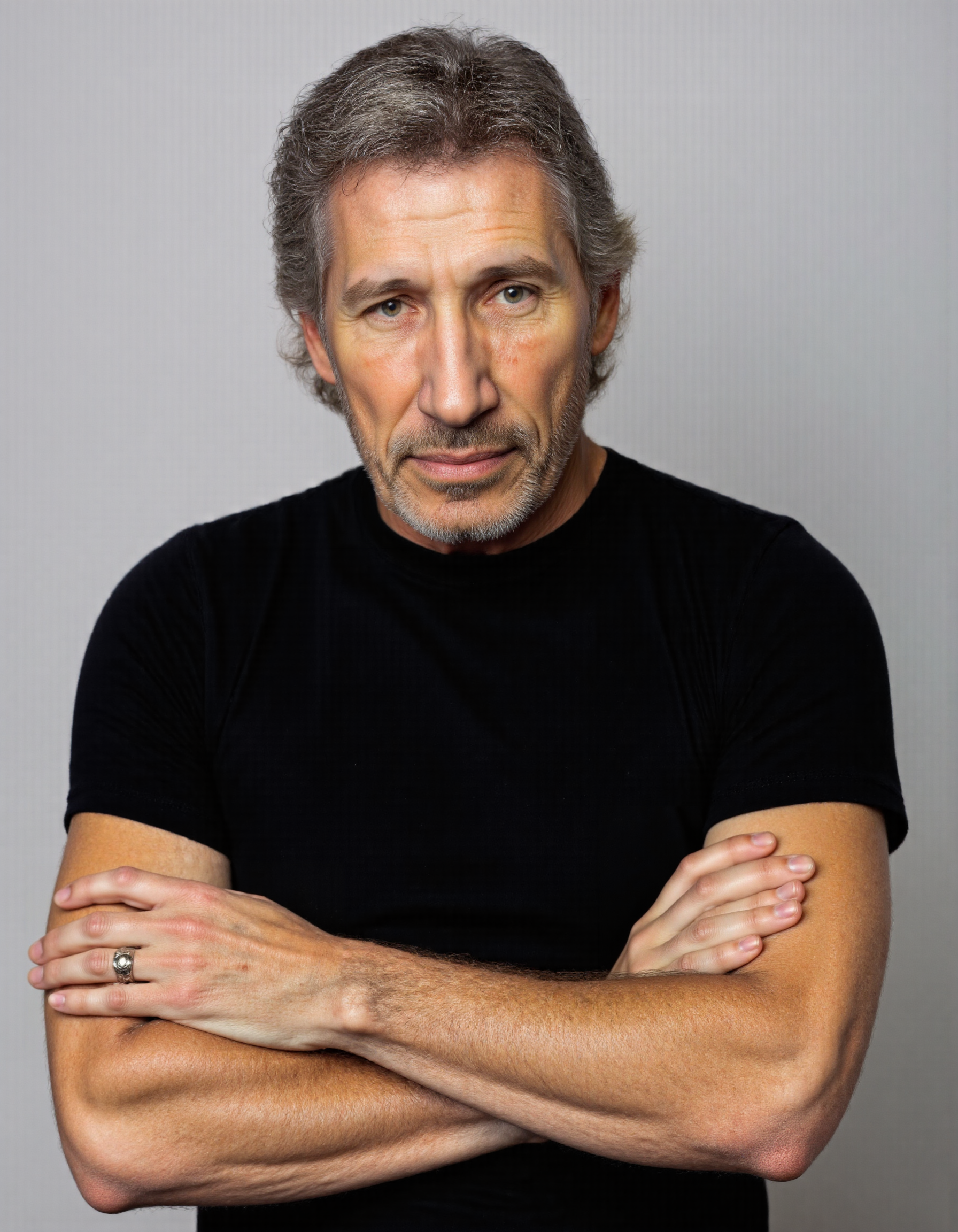 <lora:ROGER_WATERS:1>,The image is a high-resolution photograph of a middle-aged man with a relaxed and confident demeanor. He has short, wavy gray hair with a touch of dark brown, and a neatly trimmed gray beard and mustache. His skin is lightly tanned with a few visible age spots. The man is wearing a plain black t-shirt that fits snugly, accentuating his broad shoulders and muscular arms. He has his arms crossed over his chest, with his left hand resting on his right forearm, creating a casual, yet composed pose. His expression is calm and slightly smiling, with his eyes looking directly at the camera, inviting the viewer to engage with him. The background is a plain, light gray, which contrasts subtly with his dark clothing, making him the focal point of the image. The lighting is soft and even, eliminating harsh shadows and highlighting the textures of his hair and clothing. The overall mood of the photograph is warm and approachable, capturing the manâs natural, unpretentious presence.