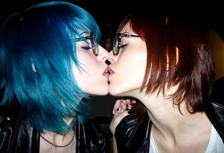 aesthetic2000s, amateur photo, two young women, kissing, intimate, blue hair, glasses, nose piercing, black leather jacket, close-up, black nail polish, affectionate, crowded frame, dramatic lighting, candid moment, pale skin, indoors, casual attire, expressive, head tilt, affectionate gesture, rebellious style, subculture, passionate scene, youth fashion, romantic interaction.,