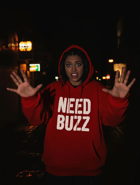 A woman standing in a dramatic, low-light urban setting, with her hands stretched out toward the viewer in a pleading gesture. She wears an oversized red hoodie featuring bold white text that reads 'NEED BUZZ'  The hoodie is slightly frayed, adding a gritty, streetwear aesthetic. Her expression is a mix of desperation and intensity, with subtle tears reflecting the dim neon lights around her. The background features a rainy alleyway, illuminated by flickering neon signs and scattered puddles reflecting the glow. The overall atmosphere is moody and cinematic, emphasizing the emotional tone of her stance and the message on her hoodie."