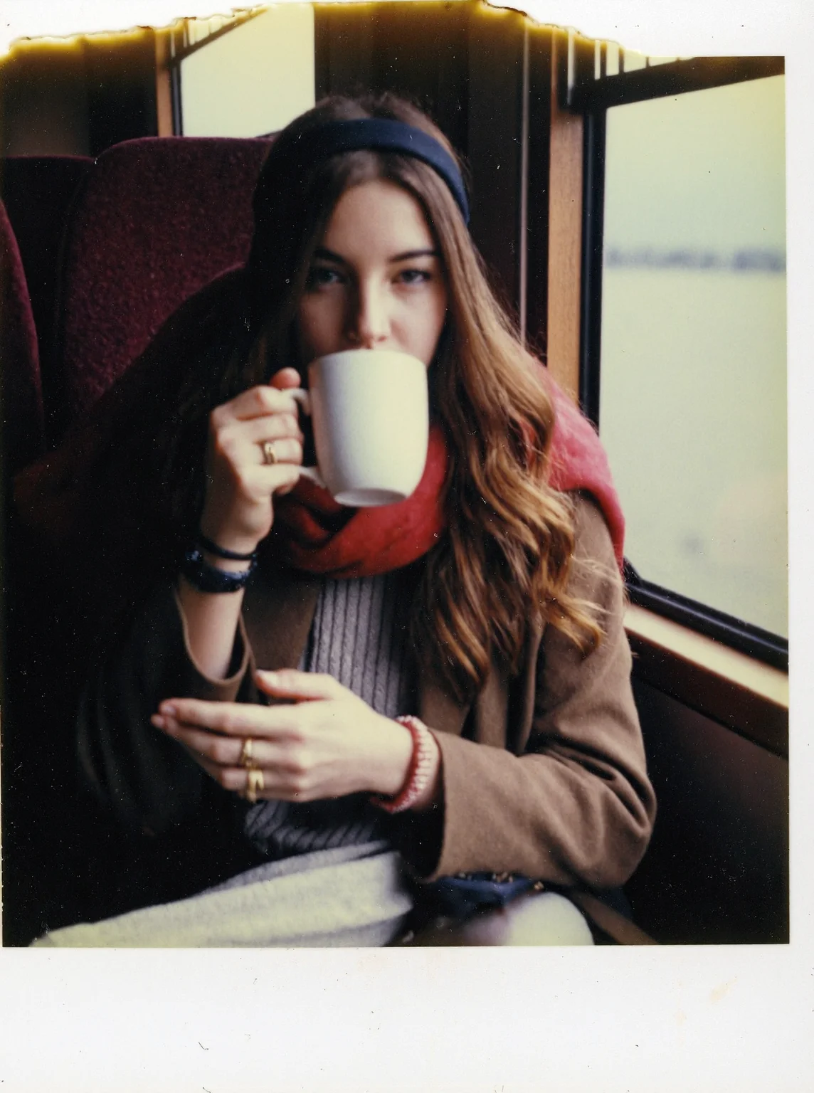 p0l2rd, Young woman seated in a train cabin, sipping from a white mug while looking directly at the camera, long wavy brown hair held back with a black headband, fair skin, wearing a vibrant red scarf over a brown coat and light-colored sweater, gold rings and accessories visible on her fingers, warm beverage likely indicated by steam or her gesture, cozy and casual posture with one knee pulled up, maroon train seat and window in the background reflecting muted outdoor scenery. prominent film grain, high contrast lighting casting dramatic shadows, grainy film-like texture, polaroid-style format with white border, distinctive burnt edge, photograph appears aged or partially developed, with the right side fading into white

