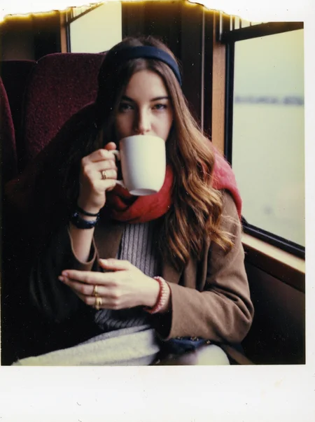 p0l2rd, Young woman seated in a train cabin, sipping from a white mug while looking directly at the camera, long wavy brown hair held back with a black headband, fair skin, wearing a vibrant red scarf over a brown coat and light-colored sweater, gold rings and accessories visible on her fingers, warm beverage likely indicated by steam or her gesture, cozy and casual posture with one knee pulled up, maroon train seat and window in the background reflecting muted outdoor scenery. prominent film grain, high contrast lighting casting dramatic shadows, grainy film-like texture, polaroid-style format with white border, distinctive burnt edge, photograph appears aged or partially developed, with the right side fading into white
