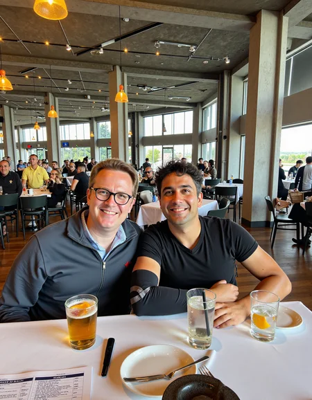 Medium shot photo of two men sitting closely together at a restaurant table, both holding drinks in a casual, friendly pose. The man on the left has light skin, short brown hair, and wears glasses with a warm smile, dressed in a dark zip-up jacket over a collared shirt. The man on the right has a medium tan skin tone, dark wavy hair, and a relaxed expression, wearing a black T-shirt with a visible arm sling. They sit side by side, leaning slightly towards each other, suggesting camaraderie, with their arms resting on the white-clothed table, which has drinks, a menu, and utensils scattered across it. The background features an expansive indoor space with high ceilings, exposed metal beams, and large windows allowing bright natural light to fill the area. The restaurant ambiance is lively, with other patrons visible in the background, creating a bustling yet comfortable atmosphere. The overall lighting is bright, diffused through the windows, giving a soft glow to the scene, emphasizing the warm, friendly interaction between the two men <lora:amateurphoto-v5-14-15-1-1:0.4>