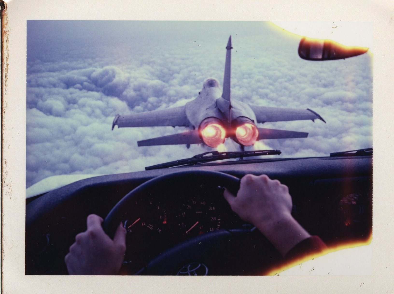 p0l2rd, Vintage photo of View from the driver’s seat of a Toyota car flying high above the clouds, young woman's hands gripping the steering wheel tightly, she has black nailpolish, dashboard gauges illuminated. Through the windshield, a fighter jet f16 is visible from the back, flying away into the distance, its twin afterburners glowing brightly with flames and leaving long, smoky vapor trails across the bright, cloud-filled sky. The sun is low on the horizon, casting warm light and subtle lens flares across the windshield. The scene captures a surreal, dynamic moment with the jet's engines prominently displayed, emphasizing its motion and power. prominent film grain, high contrast lighting casting dramatic shadows, grainy film-like texture,  polaroid-style format with white border, distinctive burnt edge, photograph appears aged or partially developed, with the right side fading into white