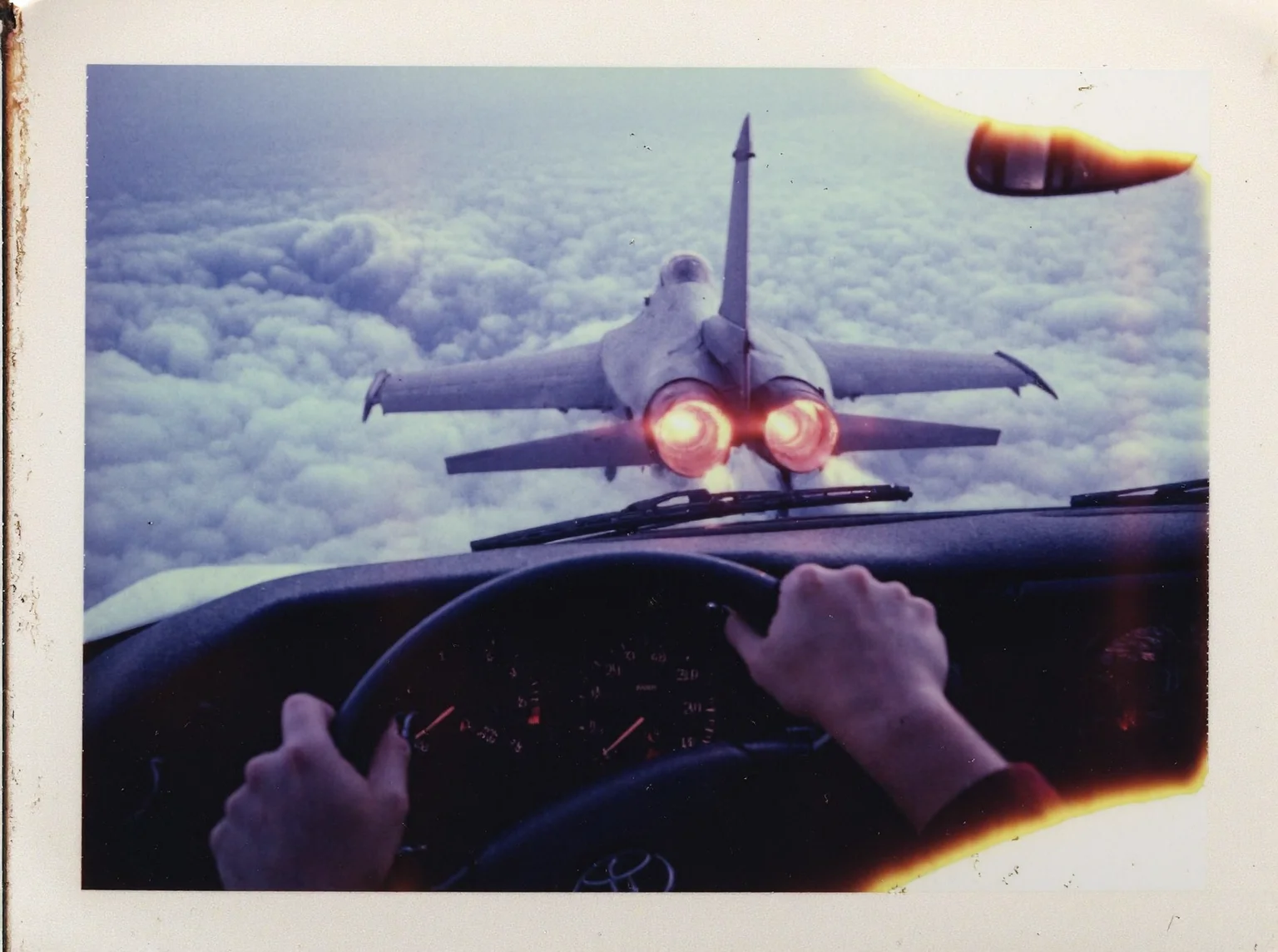 p0l2rd, Vintage photo of View from the driver’s seat of a Toyota car flying high above the clouds, young woman's hands gripping the steering wheel tightly, she has black nailpolish, dashboard gauges illuminated. Through the windshield, a fighter jet f16 is visible from the back, flying away into the distance, its twin afterburners glowing brightly with flames and leaving long, smoky vapor trails across the bright, cloud-filled sky. The sun is low on the horizon, casting warm light and subtle lens flares across the windshield. The scene captures a surreal, dynamic moment with the jet's engines prominently displayed, emphasizing its motion and power. prominent film grain, high contrast lighting casting dramatic shadows, grainy film-like texture,  polaroid-style format with white border, distinctive burnt edge, photograph appears aged or partially developed, with the right side fading into white