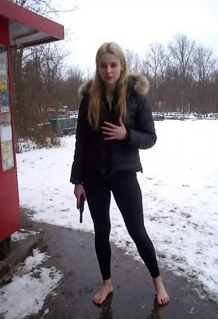 v8s,  Young woman, early 20s, casual winter attire, wearing a black puffer jacket with a faux fur hood, tight black pvc leggings, no shoes, barefoot on wet snowy ground. She stands at a food stall outdoors, holding a handgun right hand, left hand gesturing at viewer. Blonde long ponytail, natural makeup. Background shows a park, snow-covered ground, footprints, overcast weather, trees in distance, wintery atmosphere.. Amateur quality, Analog distortion, low resolution, Chromatic aberration, tape noise, Grainy texture, Frame jitter, muted colors, overexposed foreground