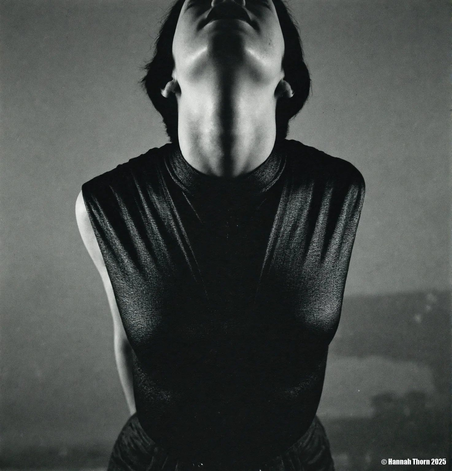 A black and white photo from a frontal camera angle, featuring a mannequin wearing a sleeveless, textured black top, positioned in the middle of the frame. the woman, who appears to be in her mid-twenties, has short, dark hair and a slim, muscular physique. she is facing the viewer, with her eyes closed and her mouth slightly open, as if she is in a state of contemplation. the background is a plain, muted grey wall, and the lighting is soft and diffused, casting shadows that accentuate the woman's face and upper body. the overall mood of the image is somber and unsettling, with a focus on the figure's facial features and clothing.<lora:_Flux\Saul_Leiter_BW.safetensors:1.0:1.0>