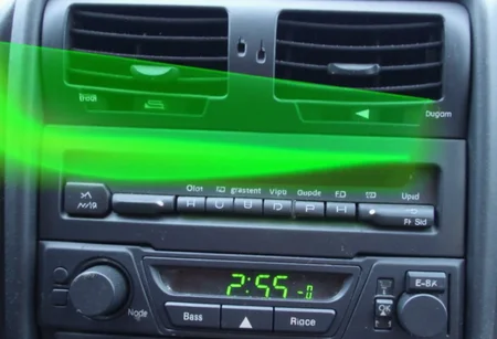 Close-up image of a car audio system console, featuring a digital display reading 2:55 in bright green digits, surrounded by various control buttons for volume, bass, treble, and radio frequency adjustment. The buttons are arranged in a clean, organized manner, characteristic of late 90s or early 2000s car stereo designs. A vivid green, ethereal streak, possibly due to light reflection, arcs across the console creating a ghost-like effect. This unusual visual element adds a surreal touch to the otherwise standard car interior. The overall image quality suggests blurred details and an amateurish quality, possibly taken in motion or with an unsteady hand.,