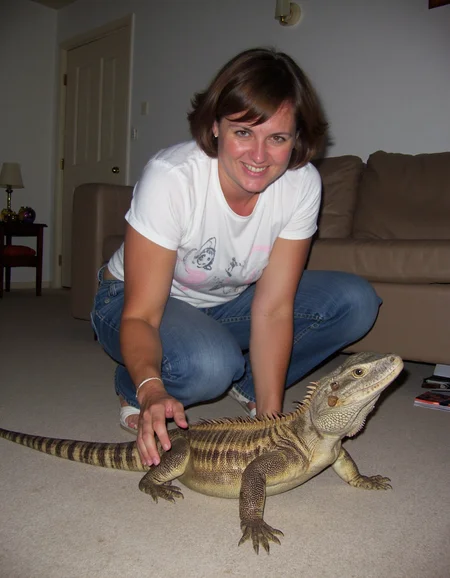 Amateur photography of a middle-aged woman with a medium build and light brown skin, wearing a casual t-shirt and jeans, playing with a Komodo dragon in her living room. The woman is crouched down, smiling, while the Komodo dragon, with its rough, scaly skin, is stretching out towards her. The living room is simple, with a plain sofa in the background, a small coffee table, and a few scattered magazines. The lighting is dim, creating shadows that obscure some details, with a bad quality sharpness to the photo, making the image look slightly overexposed and grainy, with harsh contrasts and a fine film grain effect. on flickr in 2007, 2005 blog, 2007 blog <lora:amateurphotov2-000049:0.8>