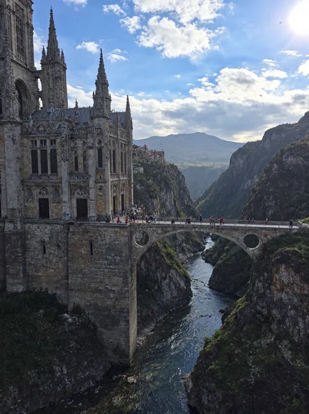 s2ms8ng. Grand cathedral-like spire, intricate Gothic carvings, pointed arches, weathered stone textures, faint moss and lichen growth on surfaces, towering archways with aged inlays, colossal structure, massive stone bridge extending across deep canyon, bridge lined with human-like figures in cloaks, detailed postures and visible shadows, lush cliffs with rugged rock textures and patches of green vegetation, meandering river below with crystal-clear water, reflections of cliffs visible on river surface, distant mountain ranges softened by haze, bright blue sky dotted with fluffy white clouds, sunlight streaming through clouds casting long shadows, scattered birds flying in the sky, mist filling the canyon, slight motion blur on cloaked figures, natural dynamic lighting, amateur quality, overhead angle