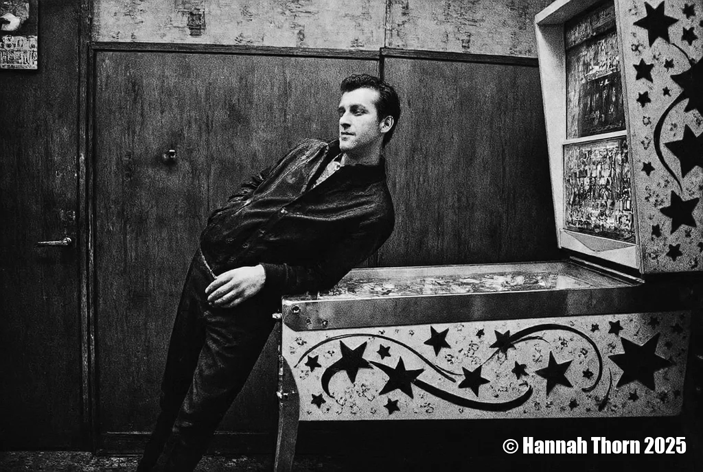 A black and white photo shoot from the side about a man standing in front of a pinball machine in a dimly lit room. the man, who appears to be in his mid-twenties, is standing on the left side of the image, leaning against the metal table with his hands on it. he is wearing a dark jacket over a pair of dark pants, and has a serious expression on his face. his hair is styled in a short, neat manner, and he is looking to the side with a slight smile. the background features a wooden door and a wall with some text on it, and the lighting is dim, casting shadows on the man's face and body. the image also shows a vintage arcade machine with a colorful design on the right side, adding to the atmosphere of the scene.<lora:_Flux\Anders_Petersen.safetensors:1.0:1.0>