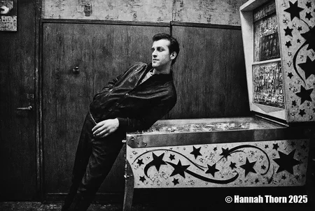 A black and white photo shoot from the side about a man standing in front of a pinball machine in a dimly lit room. the man, who appears to be in his mid-twenties, is standing on the left side of the image, leaning against the metal table with his hands on it. he is wearing a dark jacket over a pair of dark pants, and has a serious expression on his face. his hair is styled in a short, neat manner, and he is looking to the side with a slight smile. the background features a wooden door and a wall with some text on it, and the lighting is dim, casting shadows on the man's face and body. the image also shows a vintage arcade machine with a colorful design on the right side, adding to the atmosphere of the scene.<lora:_Flux\Anders_Petersen.safetensors:1.0:1.0>